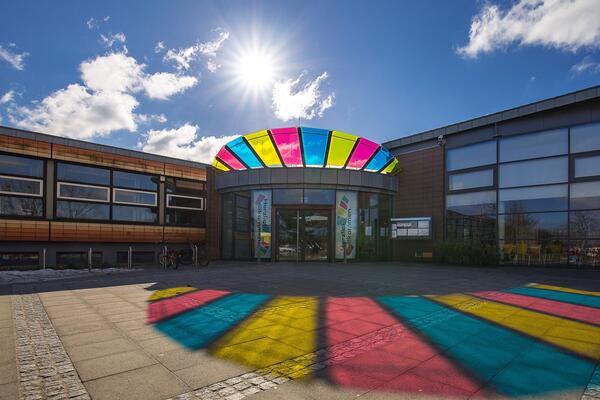 Bild vergr&ouml;&szlig;ern: Der Haupteingang der Zentralschule Harrislee spiegelt sich in den Farben (magenta, hellblau und gelb) auf dem Boden